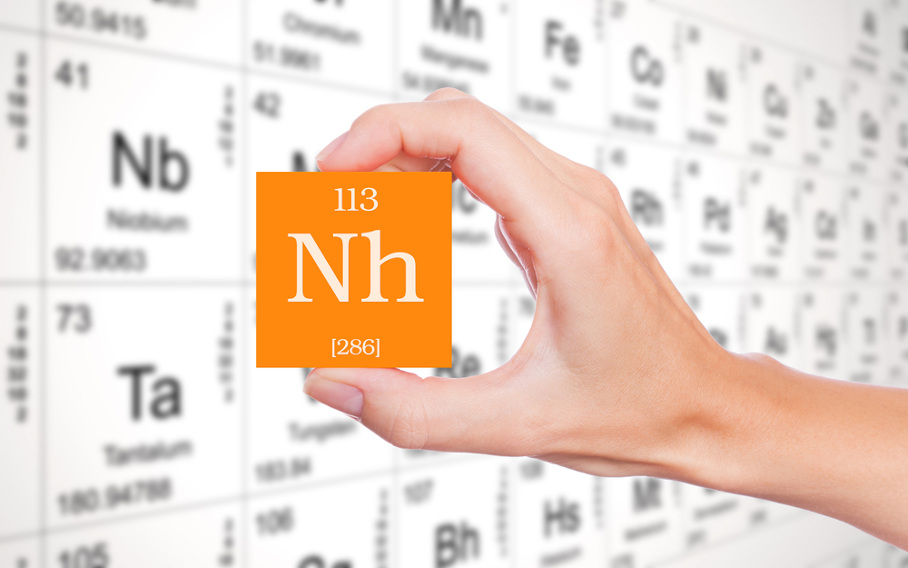 Pessoa segurando um cubo laranja, com o símbolo do nihônio, com uma Tabela Periódica branca como plano de fundo.