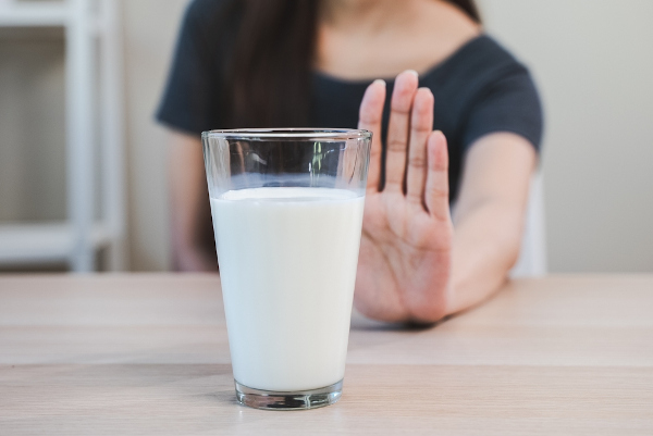 Copo cheio de leite em mesa, atrás da qual uma pessoa faz um gesto de negação a ele