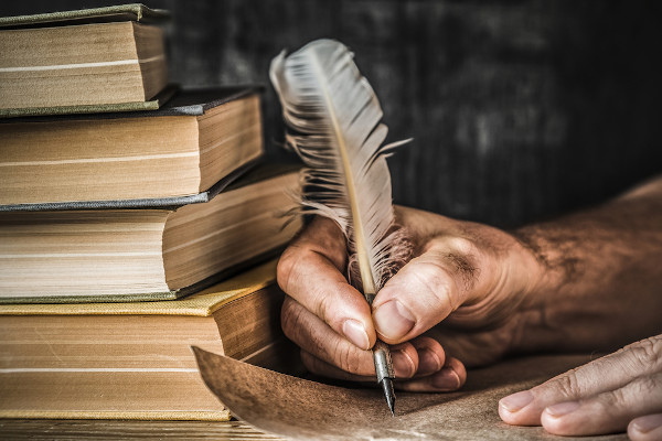 Vista aproximada da mão de uma pessoa escrevendo uma carta com uma caneta-tinteiro de pena ao lado de vários livros antigos.