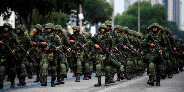 Soldados do Exército Brasileiro marchando em fileiras, uniformizados, segurando armas de fogo.
