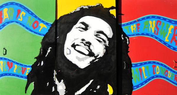 Grafite de Bob Marley em preto e branco no centro; as cores verde, amarelo e vermelho ao fundo.