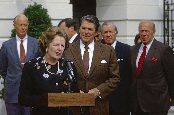 Margaret Thatcher discursando em um microfone, ao lado de Ronald Reagan; atrás deles,  outros políticos.
