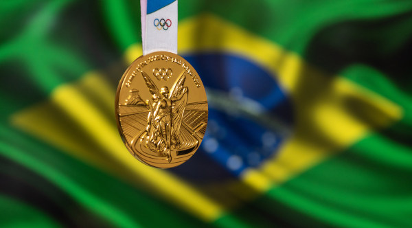 Medalha de ouro olímpica em primeiro plano, ao fundo a Bandeira do Brasil desfocada.