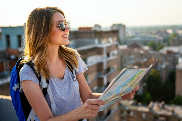 Estudante de cabelos claros e óculos escuros sorri enquanto segura mapa de viagem.
