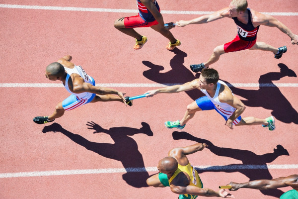 Imagem de cima de atletas passando bastões durante prova de revezamento do atletismo.