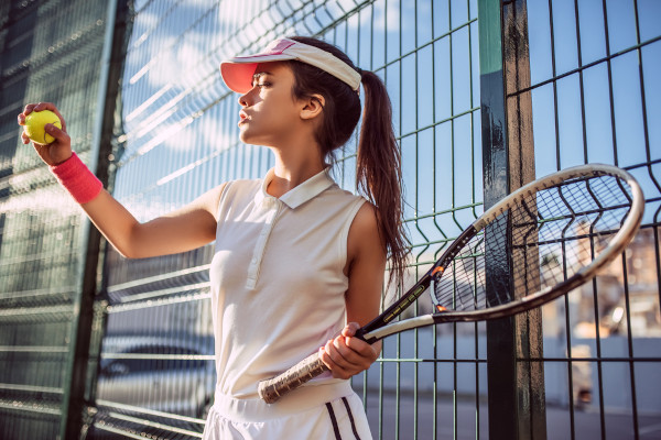 Tenista mulher usando viseira e com raquete e bola de tênis na mão.