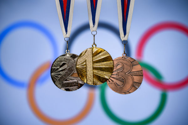 Medalhas olímpicas de ouro, de prata e de bronze, com anéis olímpicos ao fundo, uma alusão ao Jogos Olímpicos de Verão. 