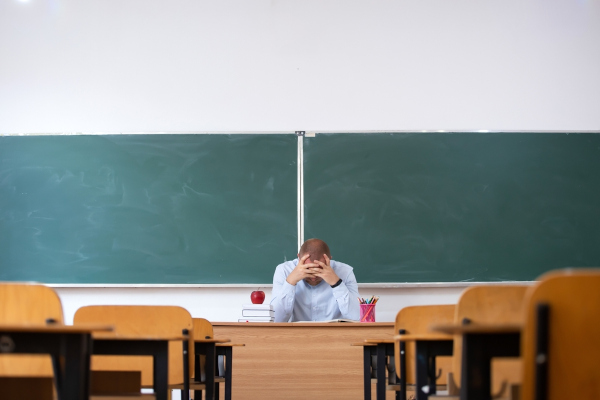 Professor sentado de cabeça baixa em uma sala de aula vazia, ilustrando as consequências da violência escolar.