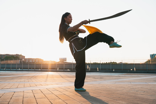 Mulher em movimento de chute ao mesmo tempo que segura espada do kung fu.
