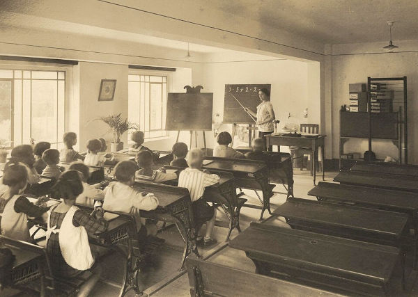 Retrato de uma sala de aula de 1926 apresenta professora no quadro-negro e alunos em carteiras escolares.