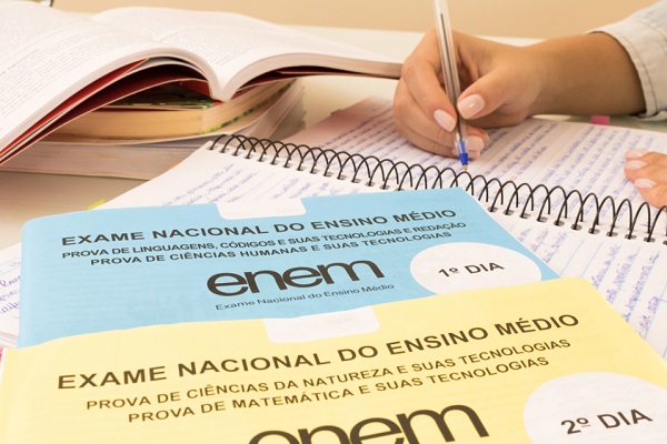 Cadernos de prova do Enem, estudante escrevendo com caneta em caderno e livros ao fundo